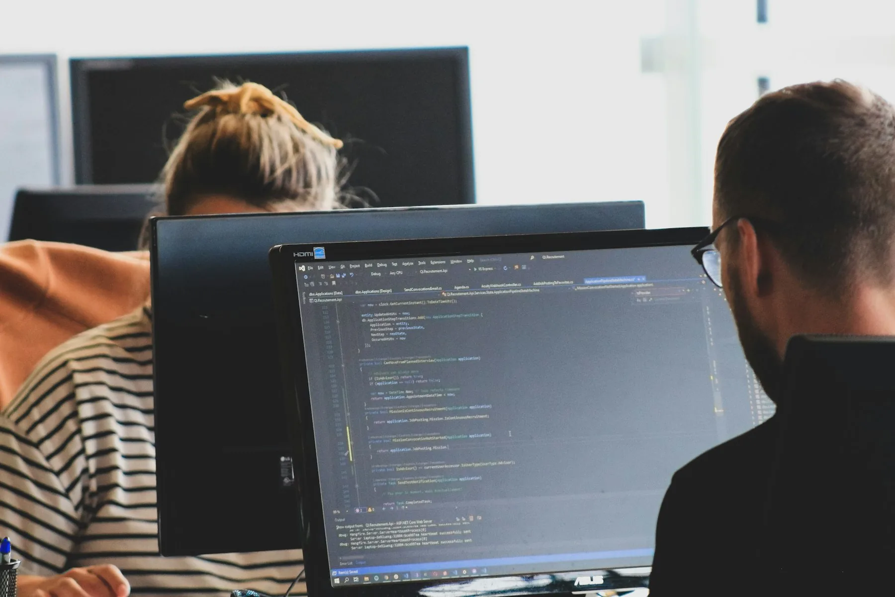 Two engineers at their monitors in a working office, one reading through code on screen.
